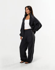 Front view of model wearing STAX. Woven Track Pants in black with open woven track jacket with contrast piping