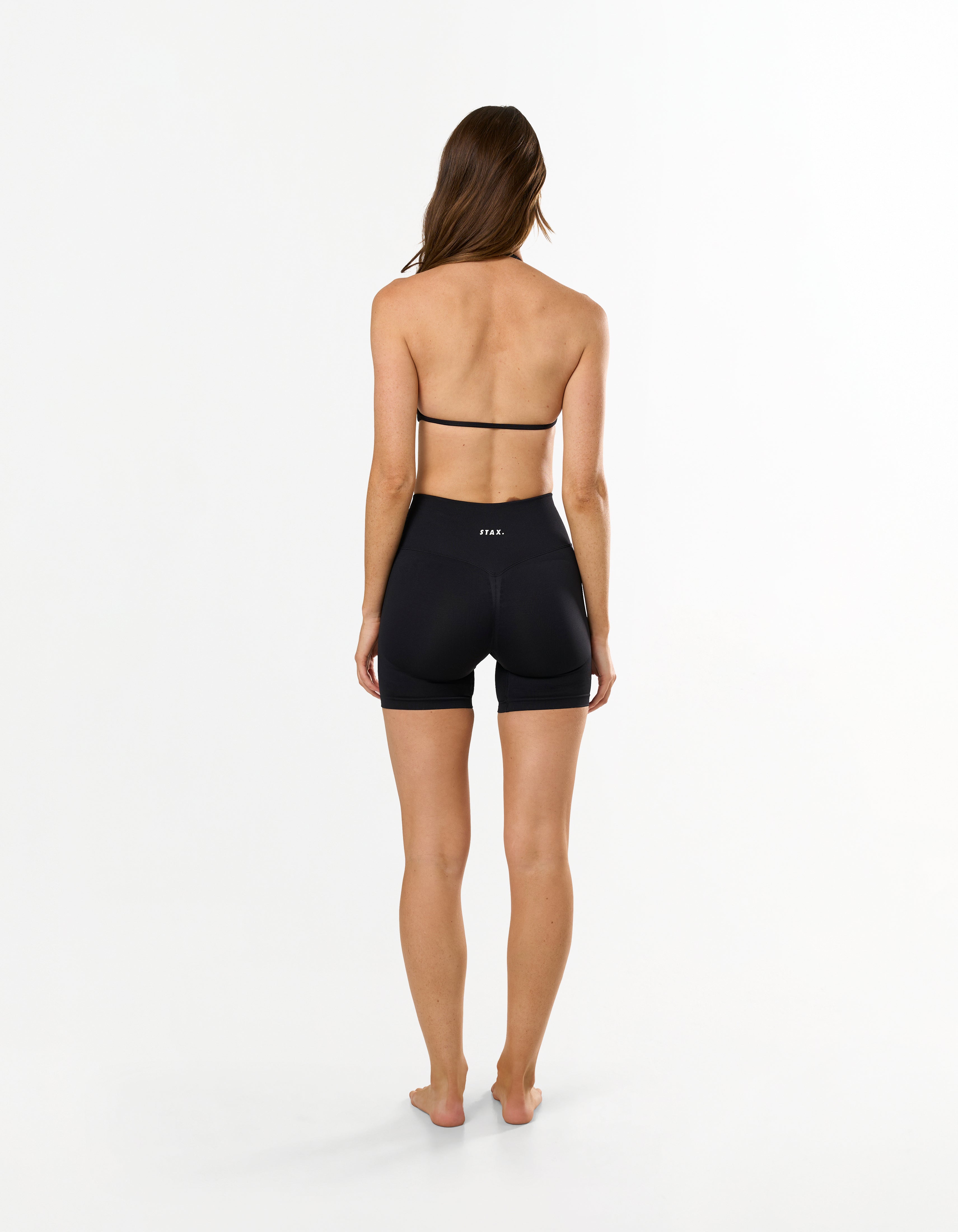 Back view of model wearing STAX. Premium Seamless Skulpt Midi Bike Shorts   in Black showing flattering invisible scrunch detail and matching strappy halter neck bralette
